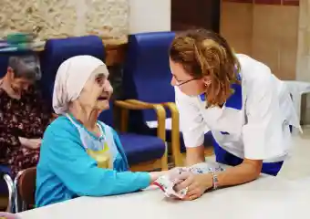 Beit Emmaus Eine Pflegekraft kümmert sich um eine alte Bewohnerin, die an einem Tisch sitzt.